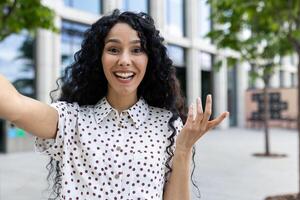 gelukkig gekruld vrouw in polka punt blouse opname en Holding camera in verspreid hand- terwijl wandelen in stad centrum. tevreden vrouw filmen vlog over nieuw gedeeld coworking voor freelancers. foto