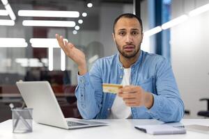 een Mens is zittend Bij een bureau met een laptop en een credit kaart in zijn hand. hij looks verward en is Holding de kaart omhoog naar zijn gezicht foto