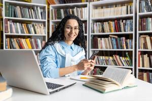 een vrolijk vrouw vervelend bril interageert met haar smartphone terwijl gezeten in een bibliotheek omringd door boeken. ze straalt uit een zin van geluk en inhoud in een academisch instelling. foto