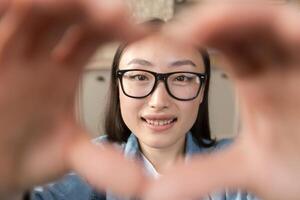 detailopname foto portret van jong mooi Aziatisch vrouw op zoek Bij camera en lachend, tiener- meisje tonen hart met handen.
