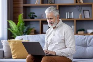 een vrolijk senior Mens met grijs haar- zit comfortabel Aan een bank, diep verloofd in gebruik makend van zijn laptop binnen een modern leven kamer versierd met binnen- planten. foto