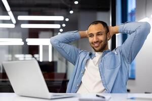 een jong Mens in een denim overhemd en gewoontjes kleding leunt terug in zijn stoel, nemen een moment naar tot rust komen in een goed verlicht kantoor opstelling. zijn houding suggereert een breken van werk, reflecterend gemak en een ontspannen atmosfeer. foto