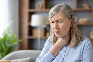 een senior vrouw verschijnen verontrust net zo ze houdt haar keel, zittend in een goed verlicht leven kamer. geeft aan mogelijk Gezondheid problemen Leuk vinden zeer keel of ongemak. foto