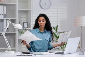 spaans vrouw op zoek verward terwijl werken met papieren en laptop in een huis kantoor instelling, illustreren werk-van-huis uitdagingen. foto