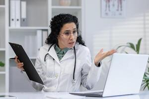 latino vrouw dokter in een kliniek hebben een telefoontje gesprek, uitleggen met hand- gebaren terwijl Holding klembord. foto