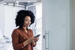 een professioneel Afrikaanse Amerikaans vrouw in een elegant polka punt blouse toepassingen haar smartphone in een helder, modern kantoor omgeving, op zoek inhoud en gefocust. foto