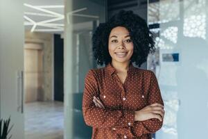 portret van gelukkig en geslaagd bedrijf vrouw, baas in overhemd glimlachen en op zoek Bij camera binnen kantoor met gekruiste armen, Afrikaanse Amerikaans vrouw met gekruld haar- in hal. foto