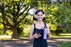 portret van een jong mooi vrouw aan het doen sport- en jogging in de park, staand in koptelefoon, Holding een telefoon en een fles van water. hij looks Bij de camera met een glimlach. foto