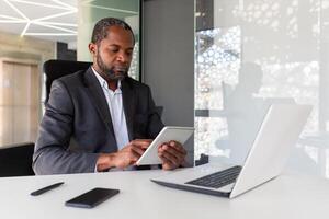 echt denken zakenman werken binnen kantoor, gebruik makend van tablet computer, Afrikaanse Amerikaans volwassen en zelfverzekerd baas gebruik makend van app Bij werkplek, lezing financieel prestatie indicatoren. foto