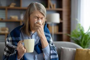 ouderen vrouw verpakt in een geruit deken niest terwijl Holding een koffie mok, zittend comfortabel Bij haar knus huis. foto