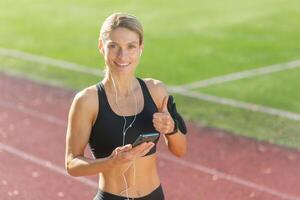 glimlachen atletisch vrouw in sportkleding met oortelefoons gebruik makend van een telefoon en geven een duimen omhoog Aan een zonnig bijhouden veld. foto