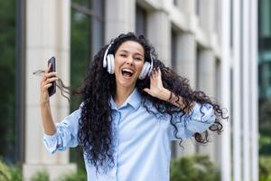 een energiek jong vrouw met lang gekruld haar, genieten van haar favoriete deuntjes met koptelefoon, Holding een smartphone buitenshuis met kantoor gebouwen in de achtergrond, overbrengen een zin vrijheid en vreugde. foto