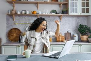 spaans bedrijf vrouw werken van een afstand van huis freelancer vieren zege en succes Holding handen omhoog op zoek Bij laptop lezing mooi zo nieuws. foto