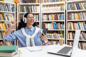 een vrolijk leerling is luisteren naar muziek- Aan koptelefoon, gebruik makend van een smartphone in een bibliotheek, omringd door boeken en een laptop. foto