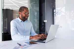 geslaagd Afrikaanse Amerikaans zakenman werken binnen kantoor met laptop, Mens glimlachen en gelukkig met prestatie resultaat Bij werkplek. foto