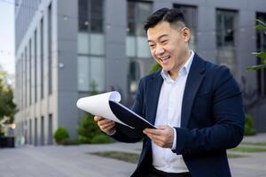 gelukkig jong Aziatisch zakenman staand in de buurt kantoor gebouw in pak op zoek Bij documenten, Holding map met papieren, tevreden met transactie en project. foto