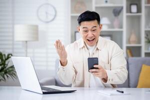 de winnaar is zittend Bij de tafel Bij huis, de Mens is gelukkig naar te ontvangen de online kennisgeving van de zege en succes Aan de telefoon, de Aziatisch Mens is Holding zijn hand- omhoog en Holding smartphone. foto