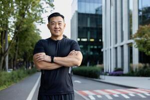 portret van een jong Aziatisch mannetje sportman staand Aan de straat van de stad in zwart sport- kleren, handen gevouwen Aan zijn borst en glimlachend op zoek Bij de camera. foto