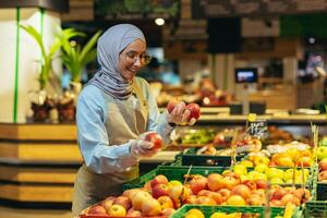 vrouw verkoper in hijab browsen en controle appels in supermarkt, vrouw in schort glimlachen Bij werk in op te slaan in fruit en groente afdeling foto