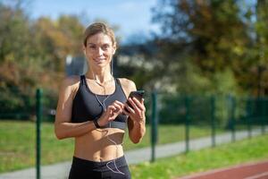 fit vrouw loper glimlachen terwijl instelling omhoog een geschiktheid app Aan haar telefoon voordat beginnend haar training Aan een zonnig spoor. foto