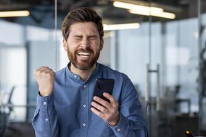 een jong professioneel in een blauw overhemd gevoel opgewonden net zo hij looks Bij zijn smartphone, vieren succes in een modern kantoor instelling. foto