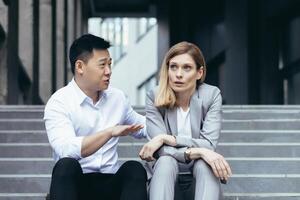 interraciaal conflict Bij werk. een jong vrouw en een jong Aziatisch Mens zijn zittend Aan de stappen in voorkant van een kantoor en ruzie maken. een Mens schreeuwt en bewijst iets naar een vrouw. de vrouw luistert moe. foto