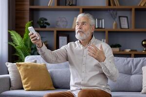 elegant senior Mens zittend Aan een grijs bank, comfortabel gebruik makend van een afgelegen controle naar bedienen de lucht conditioning in zijn stijlvol, hedendaags leven kamer. foto