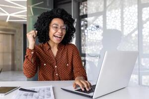 blij geslaagd tevreden Afrikaanse Amerikaans zakenvrouw vieren Super goed prestatie resultaten Bij werkplaats binnen kantoor, vrouw arbeider vieren Holding hand- omhoog glimlachen met laptop. foto