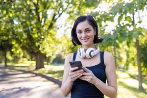 detailopname portret van een glimlachen jong vrouw aan het doen sport- in de park, gaan voor een rennen, staand in koptelefoon, Holding een telefoon en op zoek Bij de camera. foto