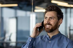 een goed gekleed Mens is verloofd in een telefoon gesprek in een strak, hedendaags kantoor omgeving, belichamen professionaliteit en bedrijf communicatie. foto