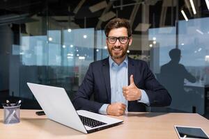 portret van volwassen geslaagd investeerder, zakenman in bedrijf pak werken binnen kantoor gebruik makend van laptop, senior mannetje baas op zoek Bij camera glimlachen en tonen duimen omhoog. foto