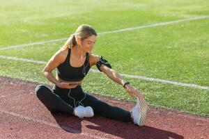 een jong vrouw atleet uitrekken haar been Aan een bijhouden veld, genieten van de ochtend- zon met hoofdtelefoons en smartwatch. foto