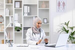 professioneel vrouw moslim dokter in hijab met tablet in een helder kliniek kantoor, het verstrekken van medisch consultaties en zorg. foto