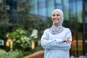 een professioneel jong vrouw vervelend een hijab vol vertrouwen glimlacht tegen een modern kantoor gebouw achtergrond, belichamen inclusiviteit en verscheidenheid in de werkplek. foto