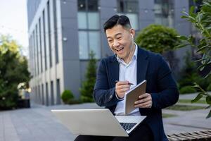 glimlachen jong mannetje Aziatisch zakenman Holding online conferentie en ontmoeting, zittend in hoofdtelefoons en pak in de buurt werk en pratend online via laptop. foto