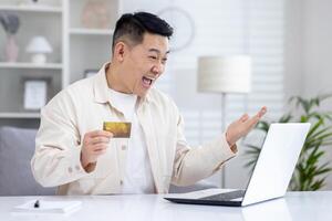 gelukkig jong Aziatisch Mens Holding credit kaart in hand- en emotioneel gelukkig op zoek Bij laptop scherm. foto