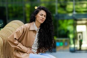 elegant Kaukasisch dame gevoel buik pijn terwijl hebben breken in buitenshuis resting Oppervlakte. van streek vrouw met gekruld haar- aanraken maag en uitdrukken pijnlijk emoties. stedelijk park. foto