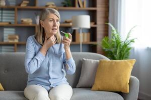 volwassen vrouw ervaren zeer keel, gebruik makend van een geneeskrachtig keel verstuiven terwijl zittend Aan haar leven kamer bank, uitdrukken ongemak. foto