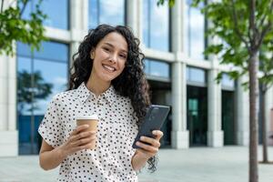 een professioneel vrouw Holding een koffie kop en smartphone buiten een hedendaags kantoor gebouw Aan een helder dag. foto