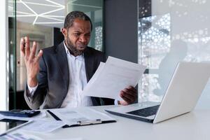 gefrustreerd en boos financier Bij werkplaats binnen kantoor, boos baas herzien facturen contracten en rapporten zittend Bij tafel met laptop, zakenman achter papier werk. foto