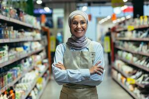 portret van een vrouw verkoper in een op te slaan tussen goederen en planken, een moslim vrouw in een schort en een hijab is glimlachen en op zoek Bij de camera, een geslaagd vrouw verkoper foto