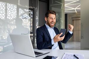 gelukkig jong zakenman zittend in de kantoor Bij de tafel, op zoek Bij de scherm van de mobiel telefoon en verheugt in succes, tonen een zege gebaar met zijn hand. foto