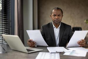 volwassen Afrikaanse Amerikaans zakenman in een pak fronst net zo hij beoordelingen belangrijk documenten, reflecterend een stressvol moment in een professioneel instelling. foto