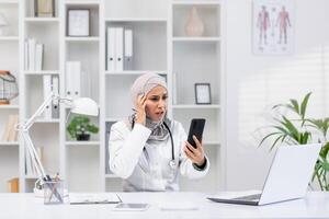 benadrukt moslim vrouw dokter met hijab op zoek bezorgd Aan een smartphone telefoontje in haar kantoor, omringd door medisch boeken en een laptop. foto