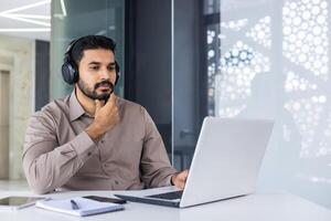 een echt denken hindoes werken binnen een kantoor met een laptop, een Mens in hoofdtelefoons luistert naar podcasts en audio boeken, een gefocust programmeur codeert nieuw software. foto