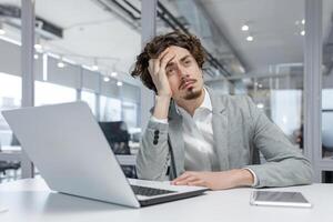 benadrukt jong zakenman met gekruld haar- nadenken oplossingen Bij zijn bureau in een hedendaags kantoor instelling, uitstralend een zin van bezorgdheid en concentratie. foto