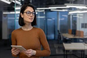 een jong professioneel vrouw in bril houdt een tablet, op zoek weg bedachtzaam in een hedendaags kantoor. foto