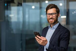 portret van geslaagd volwassen zakenman investeerder, Mens in bril en baard glimlachen en op zoek Bij camera, baas in pak Bij werk binnen kantoor Holding smartphone. foto