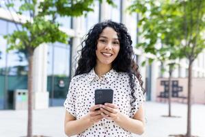 vrolijk jong vrouw met gekruld haar- sms'en Aan smartphone buitenshuis, bomen en stedelijk achtergrond, gewoontjes stijl, dag. foto