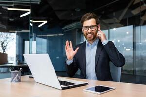 portret van een geslaagd zakenman binnen een modern kantoor, een Mens in een bedrijf pak toepassingen oortelefoons voor een telefoontje en afgelegen communicatie, een investeerder looks Bij de camera en toepassingen een laptop. foto